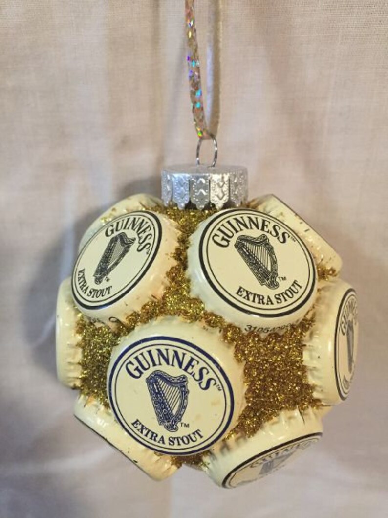 May include: A Christmas ornament made from multiple Guinness beer bottle caps, covered in gold glitter. The caps are arranged in a sphere shape and feature the Guinness logo and the words "Extra Stout".