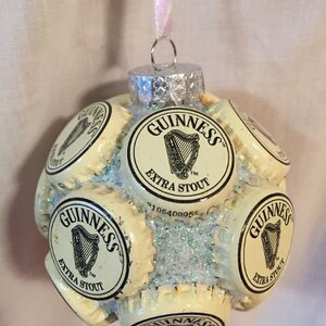 May include: A Christmas ornament made from multiple Guinness beer bottle caps. The ornament is white with black lettering and a harp logo. The ornament is covered in glitter.
