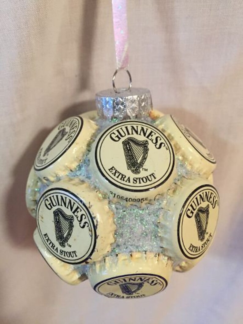 May include: A Christmas ornament made from multiple Guinness beer bottle caps. The ornament is white with black lettering and a harp logo. The ornament is covered in glitter.
