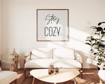 May include: A white couch with wooden arms sits in a living room with a framed print that reads "Stay Cozy" on the wall. The room has a wooden floor, a coffee table, and a potted plant.