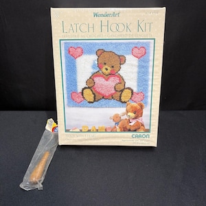 Puede incluir: Un kit de ganchillo WonderArt titulado Teddy with a Heart, que muestra un oso de peluche marrón sosteniendo un corazón rosa sobre un fondo azul y blanco. La caja muestra el diseño terminado e incluye un gancho de madera en una bolsa.