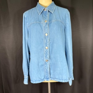 May include: Light blue denim shirt with long sleeves and a button-down front. The shirt has a classic collar and silver-colored buttons. The shirt is displayed on a mannequin against a black backdrop.