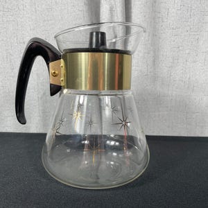 May include: A clear glass coffee pot with a gold band around the top and a black handle. The pot has a starburst design on the side.