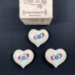 May include: Three white ceramic heart-shaped trinket boxes with floral designs. The boxes have red, blue, and green flowers painted on them. The boxes are packaged in a white box with the text "Home Interiors & Gifts, inc." and "Lasting Products Made in Mexico" printed on it.