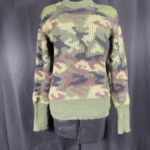 Może przedstawiać: Sweter w kamuflażu z prążkowanym okrągłym dekoltem, mankietami i dołem. Sweter ma mieszankę kolorów zielonego, brązowego i czarnego. Ramiona mają naszywki w kolorze oliwkowym. Sweter jest prezentowany na czarnym manekinie.