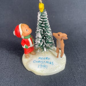 May include: A vintage Christmas ornament featuring a girl in a red coat and hat looking at a small green flocked Christmas tree with a yellow star on top. A brown deer stands next to the tree. The base of the ornament is white with the text "Merry Christmas 1981".