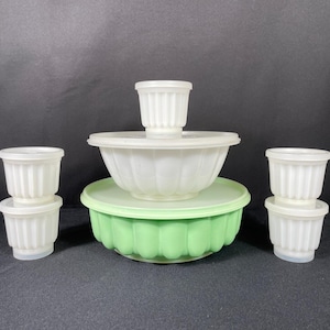 Puede incluir: Una colección de recipientes de cocina de plástico blanco y verde vintage. El conjunto incluye un cuenco grande con tapa, un cuenco más pequeño y varias tazas pequeñas acanaladas con tapas. Los recipientes están dispuestos sobre una superficie oscura.