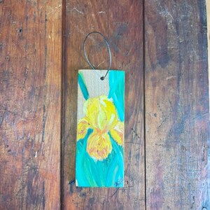 Può includere: Un fiore di iris giallo dipinto su un ornamento in legno con uno sfondo verde turchese. L'ornamento ha una corda per appenderlo.