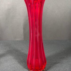 Puede incluir: Un jarrón alto de cristal rojo rubí con un diseño acanalado. El jarrón tiene un borde festoneado y una base ligeramente ensanchada con un ribete dorado. Mide aproximadamente 25 cm de altura.