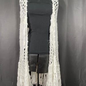 Può includere: Un gilet bianco a trama aperta con lunghe frange. Il gilet senza maniche scende sul davanti. Il design a maglia aperta è visibile su tutto il capo. Le frange pendono dal fondo del gilet.