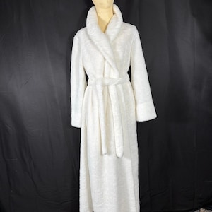 May include: A long, white, plush robe with a shawl collar and a matching belt. The robe has long sleeves and is displayed on a mannequin against a black backdrop. The robe is made of a soft, textured material.