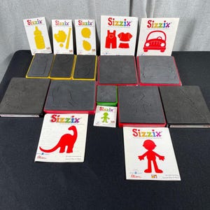 Könnte beinhalten: Eine Sammlung von Sizzix Stanzmaschinen und Stanzformen. Die Stanzformen zeigen verschiedene Formen, darunter ein Auto, einen Dinosaurier und eine Person. Die Verpackung ist weiß mit dem Sizzix-Logo und rot oder gelb gestanzten Formen.
