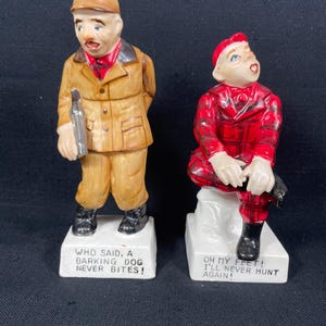 May include: Two ceramic figurines of men dressed in hunting attire. The man on the left is holding a gun and has a surprised expression on his face. The man on the right is sitting down with his legs crossed and has a shocked expression on his face. The figurine on the left has the text "WHO SAID, A BARKING DOG NEVER BITES!" on the base. The figurine on the right has the text "OH MY FEET! I'LL NEVER HUNT AGAIN!" on the base.