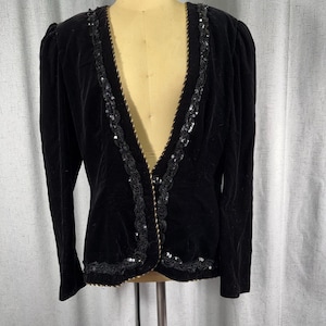 May include: A black velvet blazer featuring long sleeves, a deep V-neckline, and a single front button closure. The edges of the neckline and the hem are decorated with black lace trim, shimmering black sequins, and a thin gold-toned braided cord.