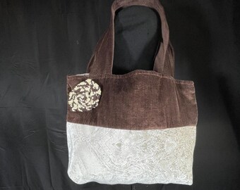 Pode incluir: Uma bolsa tote de veludo cotelê marrom com uma base de tecido branco. A bolsa tem um coração de crochê branco e marrom preso na frente.