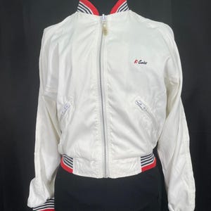 May include: White K-Series jacket with a full zipper, red and navy blue striped accents on the collar, cuffs, and waist. The jacket has two zippered pockets and the K-Series logo embroidered on the chest. The jacket is displayed on a black mannequin.