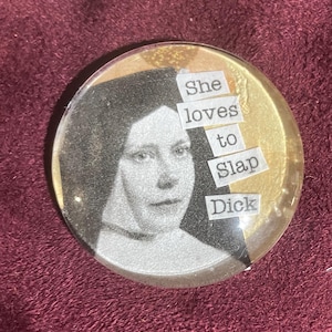 Puede incluir: Botón redondo y transparente con un retrato vintage en blanco y negro de una mujer. El botón tiene purpurina dorada alrededor del retrato y el texto dice "She loves to slap Dick."