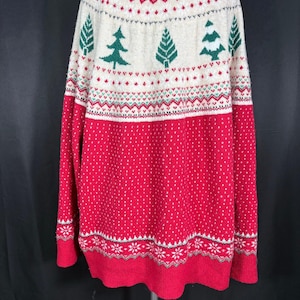 May include: A long-sleeve knit sweater featuring a red lower section with white speckles and a cream-colored upper section. The design includes green pine tree motifs, red and green geometric patterns, and white snowflake borders at the hem.