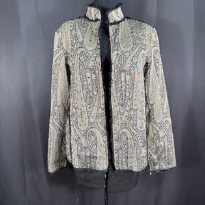 May include: A long-sleeved jacket featuring a black and beige paisley pattern throughout. The garment has a high collar, an open front design, and visible frayed black trim along the edges, collar, and front opening, creating a textured appearance.