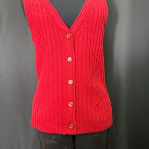 Könnte beinhalten: Eine rote, ärmellose Strickweste mit Knopfleiste und V-Ausschnitt. Die Weste hat ein Rippenstrickmuster mit dekorativen Nähten an den Seiten und goldfarbenen Knöpfen. Das Kleidungsstück wird auf einer schwarzen Schaufensterpuppe präsentiert.