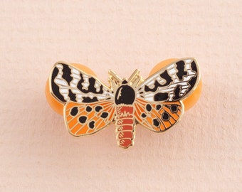 Puede incluir: Un pin de esmalte dorado con una mariposa negra, blanca y naranja con manchas negras. La mariposa está sobre un fondo rosa claro.
