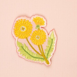May include: Embroidered patch featuring three yellow dandelion flowers with green leaves and stems. The patch has a pink border and is set against a light pink background. The flowers have detailed stitching.