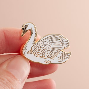 Może przedstawiać: Złota szpilka emaliowana przedstawiająca białego łabędzia z czarnymi oczami. Łabędź jest przedstawiony w eleganckiej pozie z rozłożonymi skrzydłami.