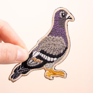 Puede incluir: Parche bordado de una paloma en tonos grises, morados y beige. El ave presenta un diseño detallado con detalles de alas en blanco y negro y patas amarillas. El parche se sostiene sobre un fondo blanco roto.