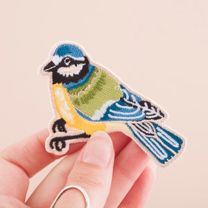 Puede incluir: Un parche bordado con un pájaro azul con detalles amarillos, verdes y azules. El pájaro está posado en una rama con las alas extendidas.