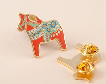 May include: A red and gold enamel pin of a traditional Swedish Dala horse with floral details. The pin has a gold metal back with a safety clasp.
