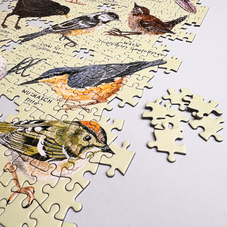 May include: A completed jigsaw puzzle with a light yellow background featuring illustrations of different birds. The puzzle pieces are arranged to show a wren, a coal tit, a nuthatch, and a goldcrest. The puzzle is missing a few pieces.