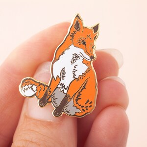 May include: A gold-tone enamel pin featuring a stylized orange fox sitting with its tail curled behind it.