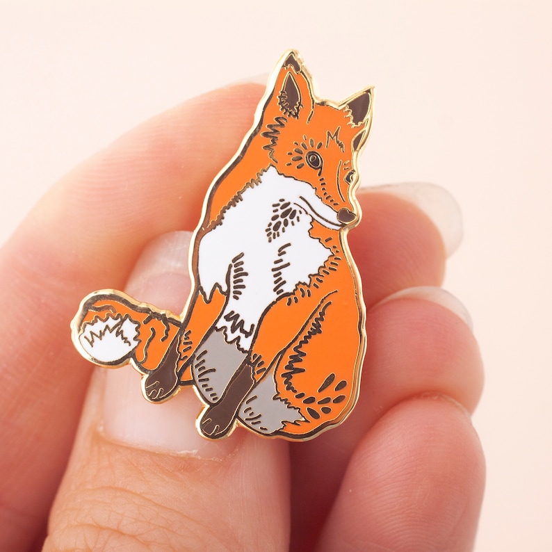 May include: A gold-tone enamel pin featuring a stylized orange fox sitting with its tail curled behind it.