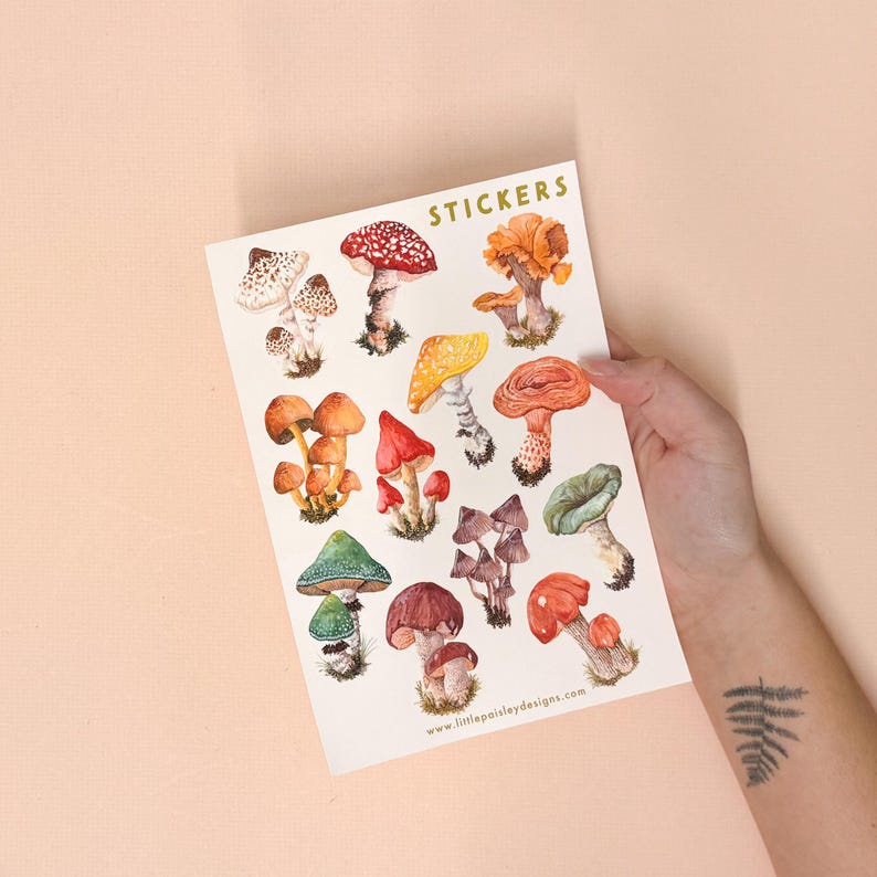 May include: A sheet of stickers featuring a variety of colorful mushroom illustrations. The stickers are arranged in a grid pattern and include different types of mushrooms, such as red and white spotted mushrooms, yellow mushrooms, and brown mushrooms. The sheet is labeled "STICKERS" at the top and includes the website "www.littlepaisleydesigns.com" at the bottom.
