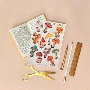 May include: A sheet of stickers featuring colorful illustrations of various mushrooms. The stickers are arranged in rows and columns on a white background. The text "STICKERS" is printed in gold lettering at the top of the sheet.