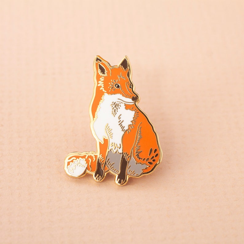 May include: A gold-tone enamel pin featuring a red fox with a white chest and a curled tail. The fox is sitting on its haunches and looking to the right.