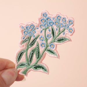 May include: A pink embroidered patch featuring a cluster of blue forget-me-not flowers with green stems and leaves.