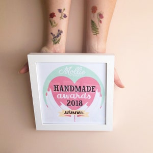 Puede incluir: Una impresi&oacute;n enmarcada en blanco con un coraz&oacute;n rosa y el texto "Mollie Makes Handmade awards 2018 winner" sobre un fondo azul claro.