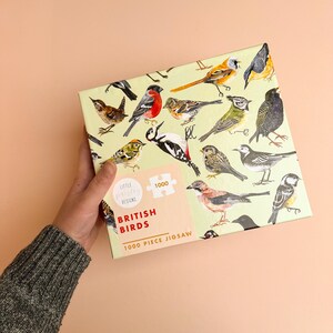 May include: A 1000 piece jigsaw puzzle featuring a variety of British birds on a light green background. The puzzle box has a light green background with a white border and the text "British Birds 1000 Piece Jigsaw" in red and black lettering. The puzzle box also features a small logo for "Little Pixel Designs".