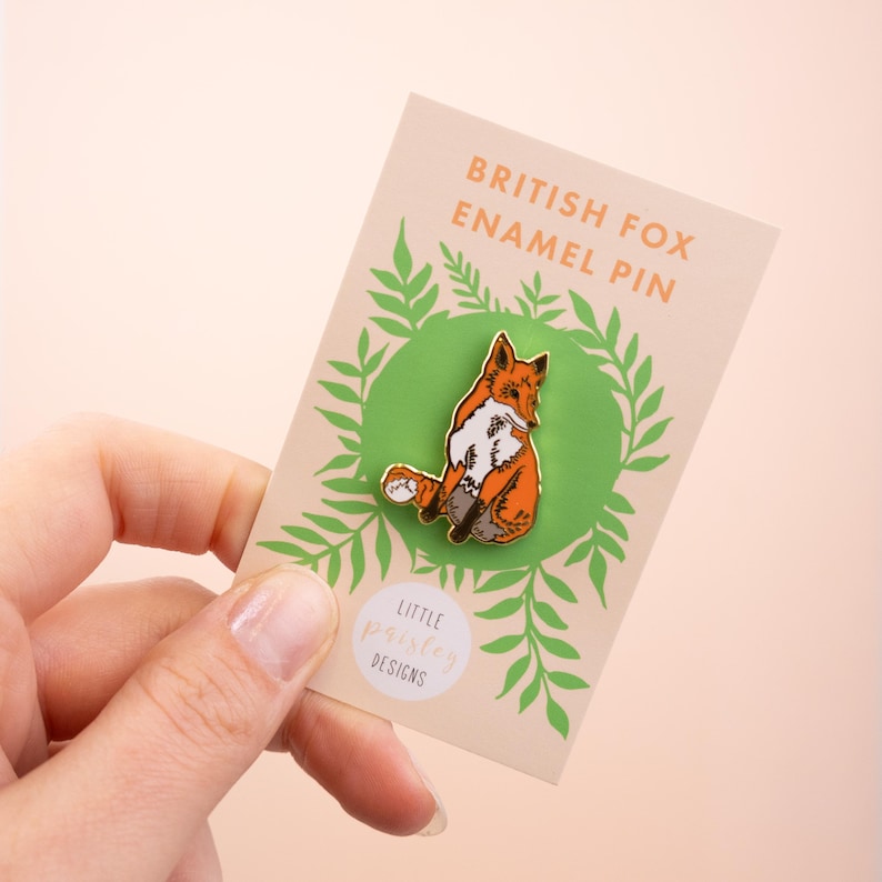 May include: A gold enamel pin featuring a red fox sitting on a green background with a leafy border. The pin says "British Fox Enamel Pin" and "Little Paisley Designs".