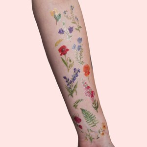 Puede incluir: Un brazo con una variedad de tatuajes temporales florales de colores. Los tatuajes incluyen amapolas, nomeolvides y helechos.