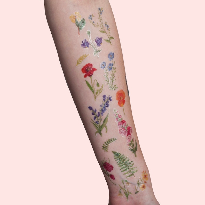 Puede incluir: Un brazo con una variedad de tatuajes temporales florales de colores. Los tatuajes incluyen amapolas, nomeolvides y helechos.