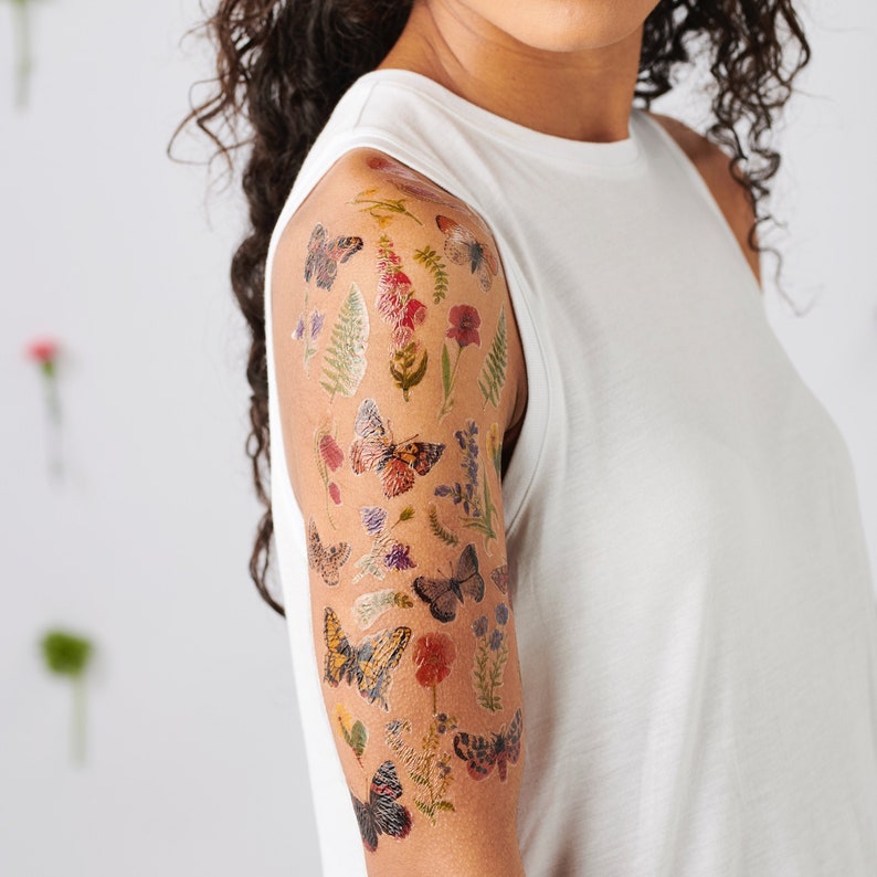 Puede incluir: Un brazo con tatuajes temporales de mariposas y flores. Los tatuajes son en tonos de rojo, amarillo, verde y azul.