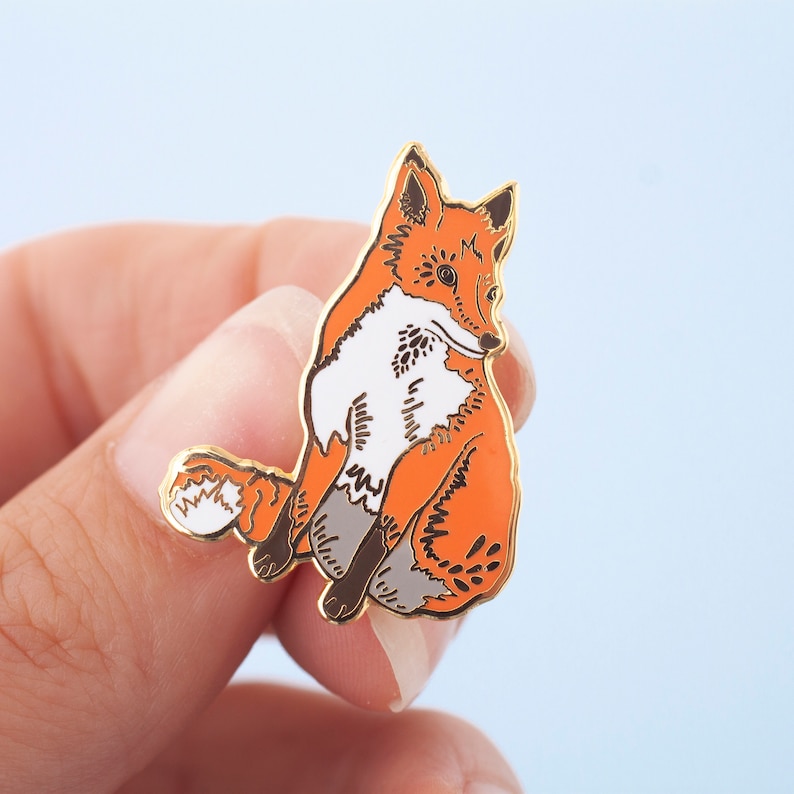 May include: A gold-tone enamel pin featuring a red fox with white markings sitting on its haunches. The fox has a detailed design with black eyes and a black nose.
