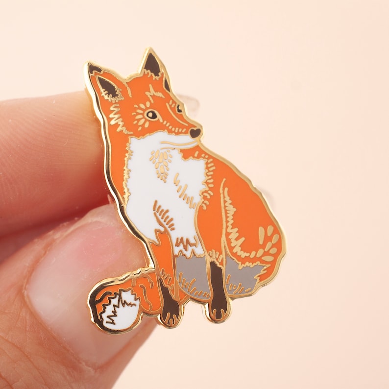 May include: A gold-tone enamel pin featuring a detailed illustration of a red fox with white markings. The fox is sitting with its tail curled around its body.