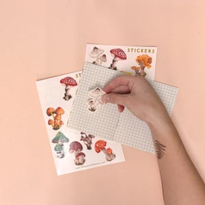 May include: A sheet of mushroom stickers with a variety of mushroom illustrations. The stickers are on a white background with the text "STICKERS" in gold lettering. A hand is holding a mushroom sticker.