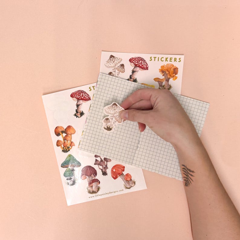 May include: A sheet of mushroom stickers with a variety of mushroom illustrations. The stickers are on a white background with the text "STICKERS" in gold lettering. A hand is holding a mushroom sticker.