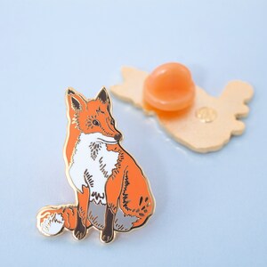 May include: A gold-tone enamel pin featuring a red fox with white markings sitting on its haunches. The fox has a curled tail and is looking to the left. The pin has a gold-tone back with a safety pin closure.