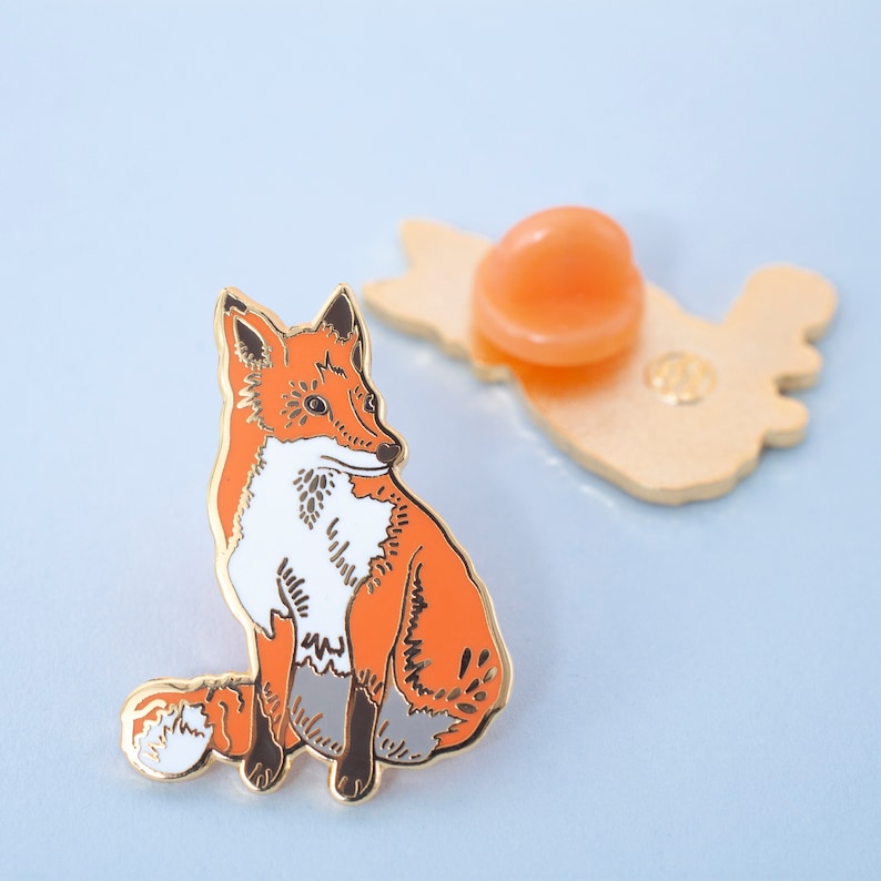 May include: A gold-tone enamel pin featuring a red fox with white markings sitting on its haunches. The fox has a curled tail and is looking to the left. The pin has a gold-tone back with a safety pin closure.