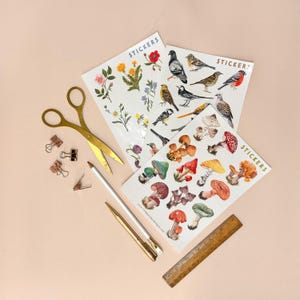 May include: Three sheets of stickers featuring illustrations of mushrooms, birds, and flowers. The stickers are on a light pink background with gold scissors, paper clips, and pencils.